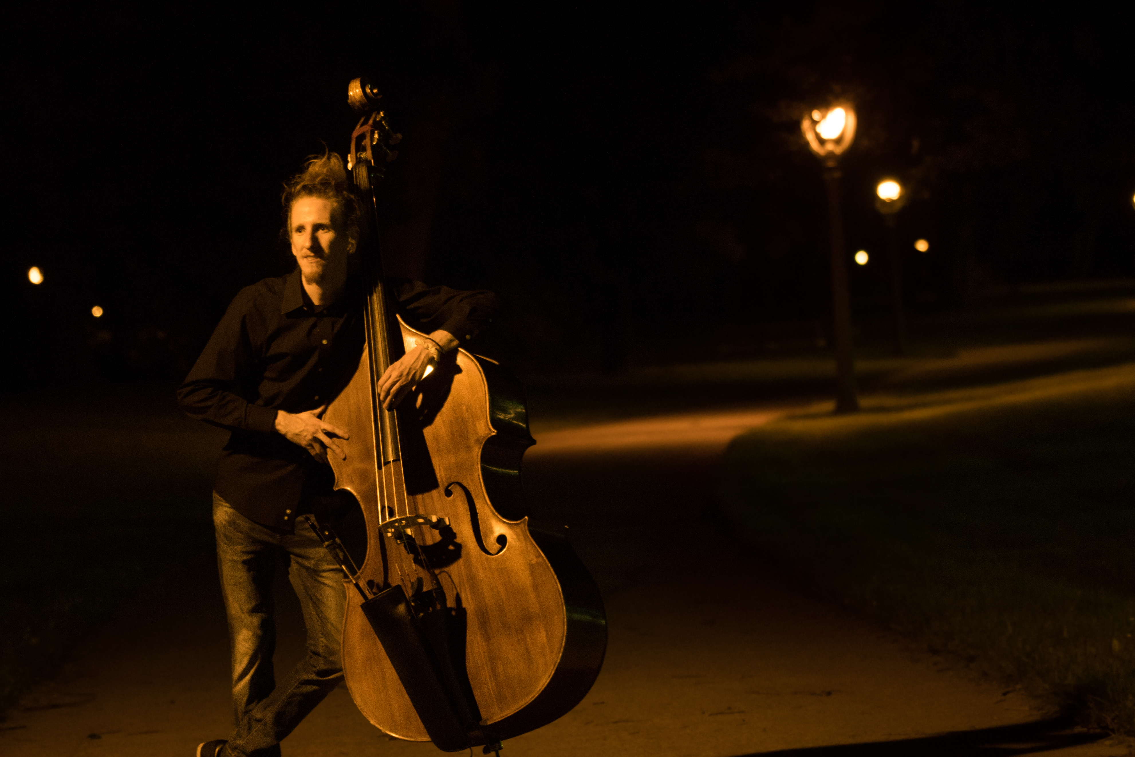 Photo of Sam posing with his upright bass, taken at night, trees and street lamps 
								in the background.